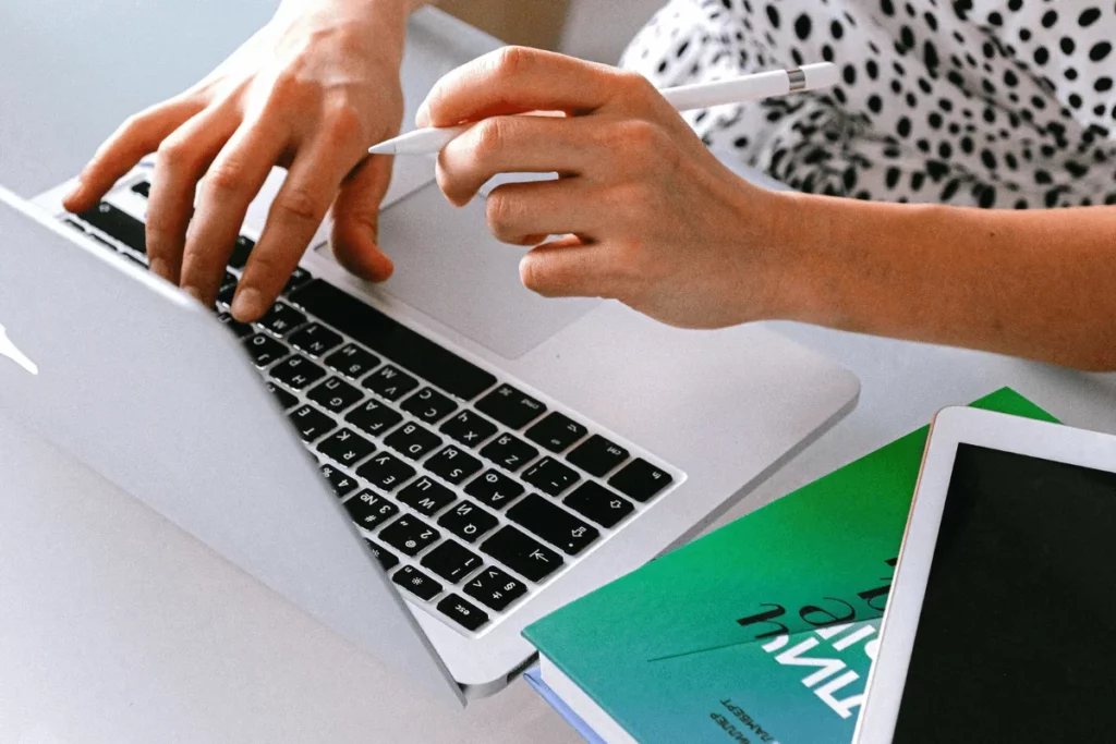 Individual typing on a laptop, with a pen in hand and a green book on the desk.