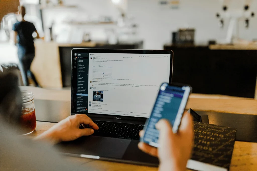 Person using a laptop and smartphone to communicate on a messaging app in a cozy workspace.