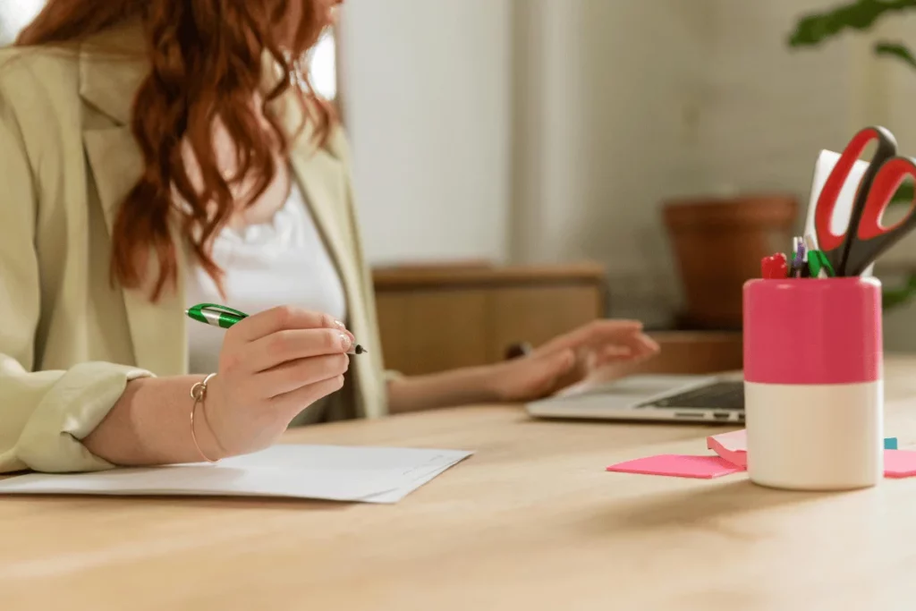 A focused woman in a light jacket holding a pen over papers at an office desk.
