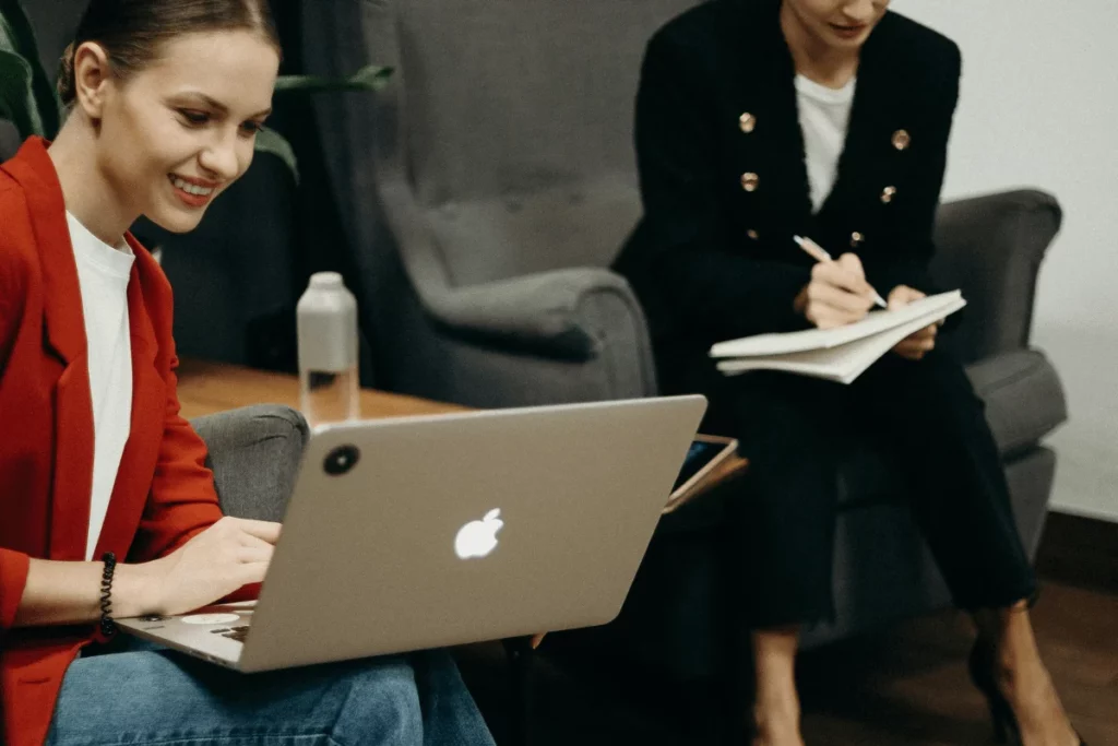 Two women in a meeting, one using a laptop and the other writing in a notebook.