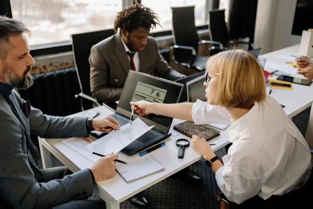 A diverse team collaborates around a table with laptops and documents, engaging in focused discussion. The atmosphere is professional and serious.