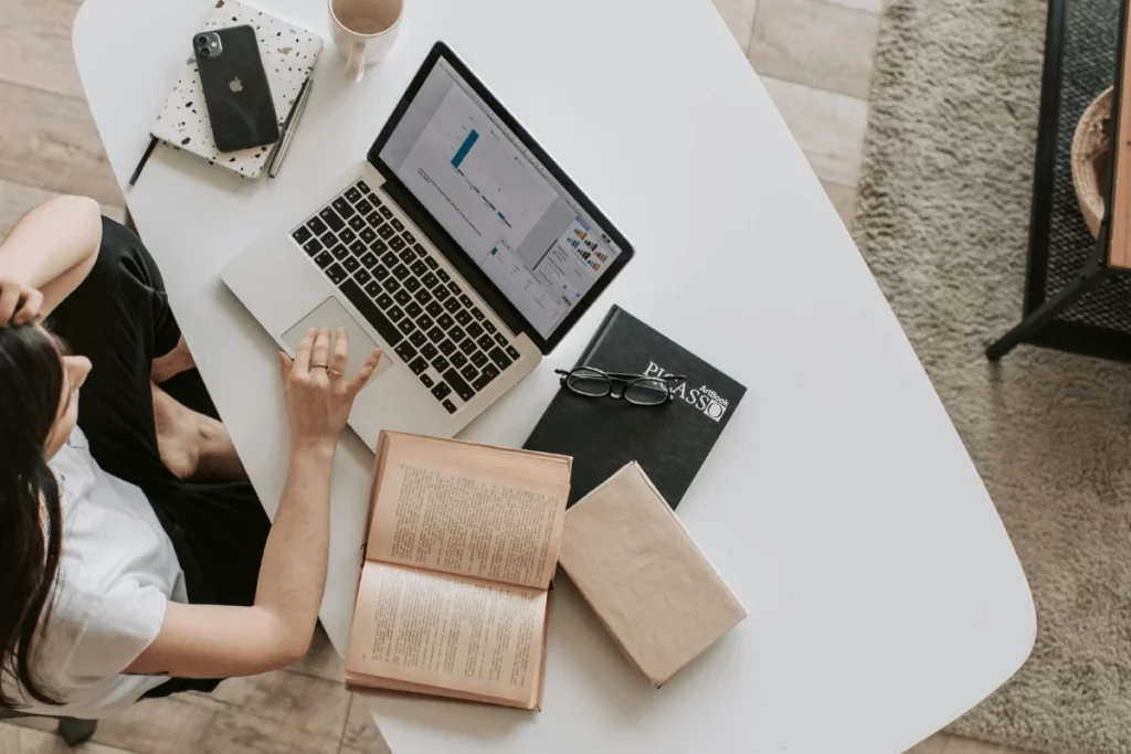 Overhead view of a person at a white table working on a laptop near an open book.