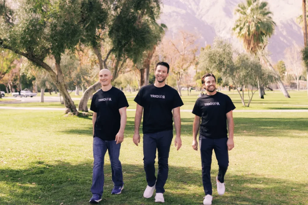 Three men wearing matching branded T-shirts walking in a sunny park with trees and mountains in the background.