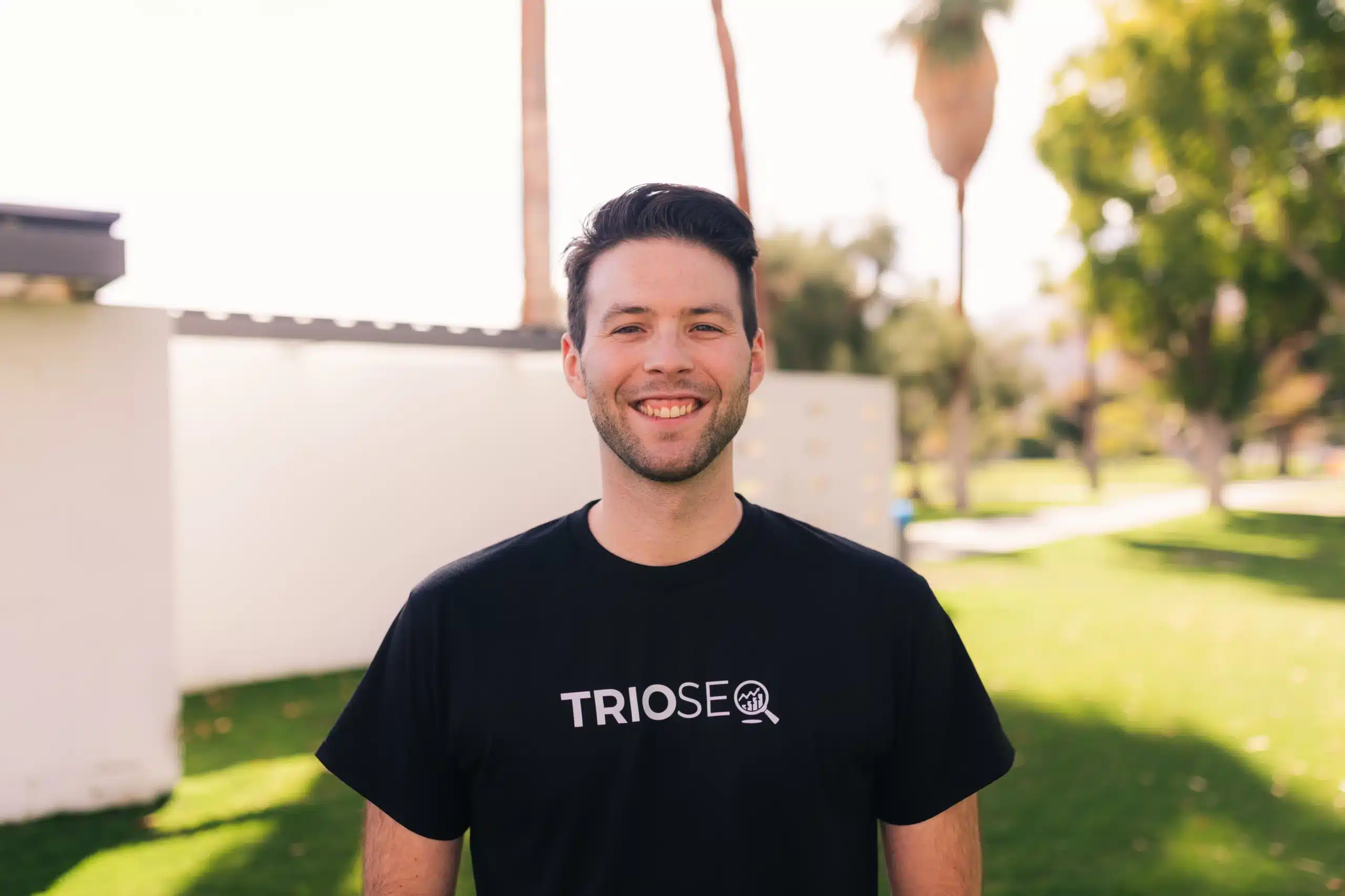 Steven Schneider stands outdoors wearing a black t-shirt with "TRIOSEO" printed on it, surrounded by greenery and soft sunlight.